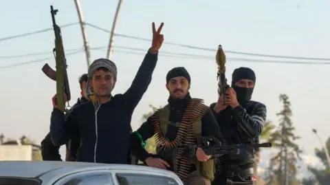 Getty Images Three young men holding weapons pose on the back of a pick up truck. One has a RPG, another is holding a heavy machine gun. The man at the front holds a AK47-style assault rifle and is raising his fingers in salute.