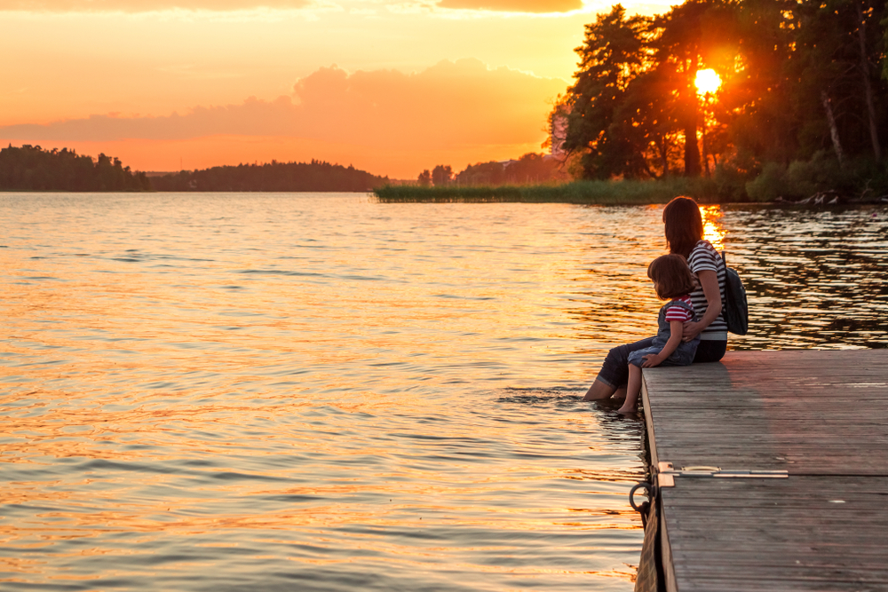 A woman and a young girl sit side by side on a dock with their feet in the water while they watch the sunset. 