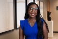 Une femme noire aux cheveux longs, portant des lunettes, une robe bleu roi et un collier en argent, sourit à la caméra.