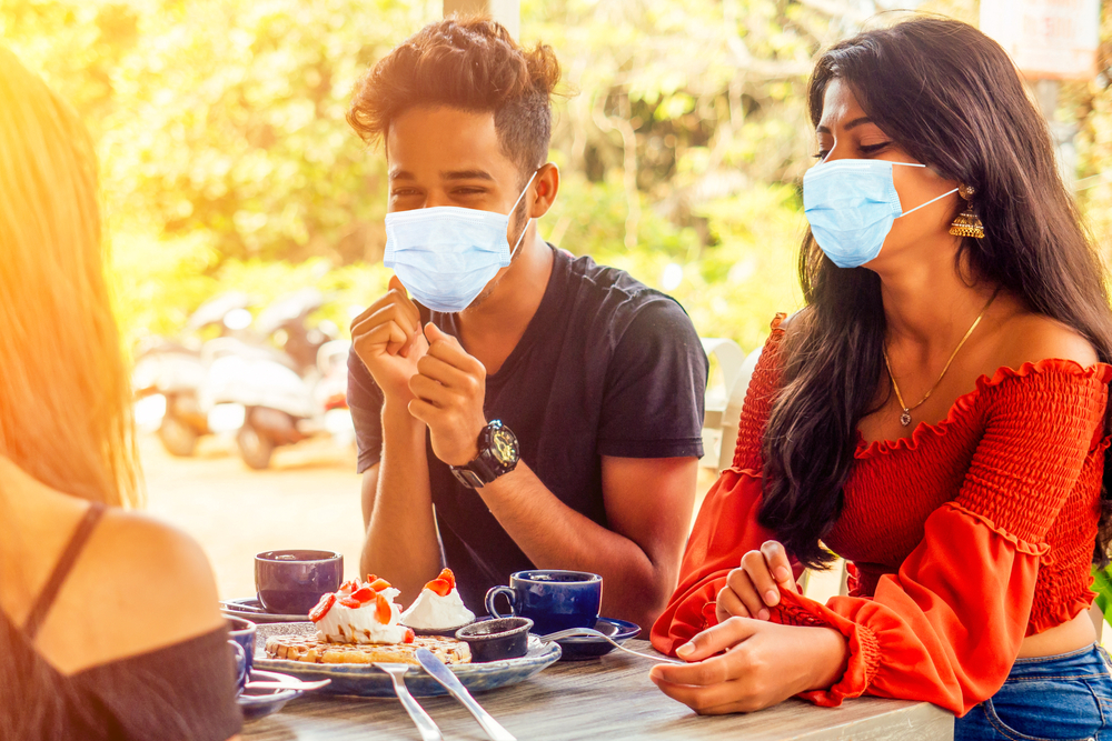 A group of young people wearing masks enjoy a get-together on a patio on a summer day. 