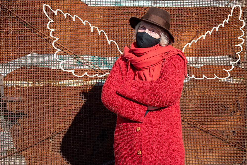 An older white woman wearing a mask and a red coat stands in front of a wall with angel wings painted on them, so it looks like the wings are hers. 