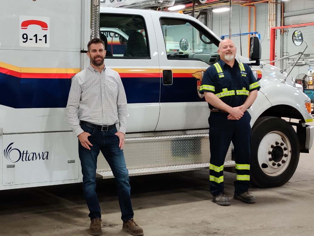 A bearded man in a button-down shirt, jeans and boots and a man with a goatee wearing a blue uniform with reflective paneling stand in front of an ambulance in a garage. 