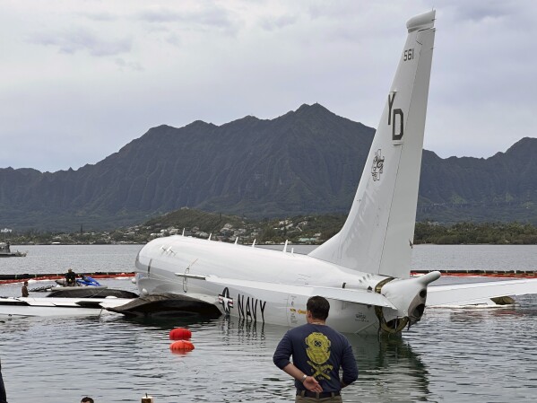 Contractors place inflatable bags under a U.S. Navy P-8A in Kaneohe Bay, Hawaii, Friday, Dec. 1, 2023, so they can float the aircraft over the water and onto land. The Navy plans to use inflatable cylinders to lift the jet off a coral reef and then roll it over to a runway to remove the plane from the ocean where it crashed the week before. (AP Photo/Audrey McAvoy)