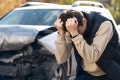 Distressed young man clutching his head with a crashed car behind him/Un jeune homme accablé agrippe sa tête tandis qu’une voiture accidentée se trouve derrière lui.