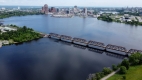 Bird’s eye view of the Chief William Commanda Bridge over the water with tree-lined pathways and City buildings, including Parliament, in the background.