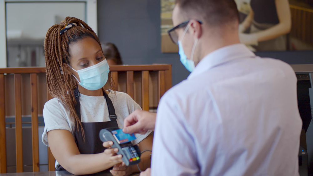 A young black woman wearing a mask accepts payment from a customer at a restaurant. 