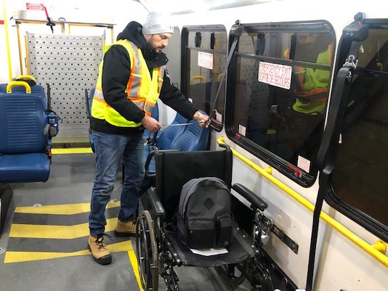 A male trainee in a grey hat and a safety vest practices securing a wheelchair on a Para Transpo bus. 