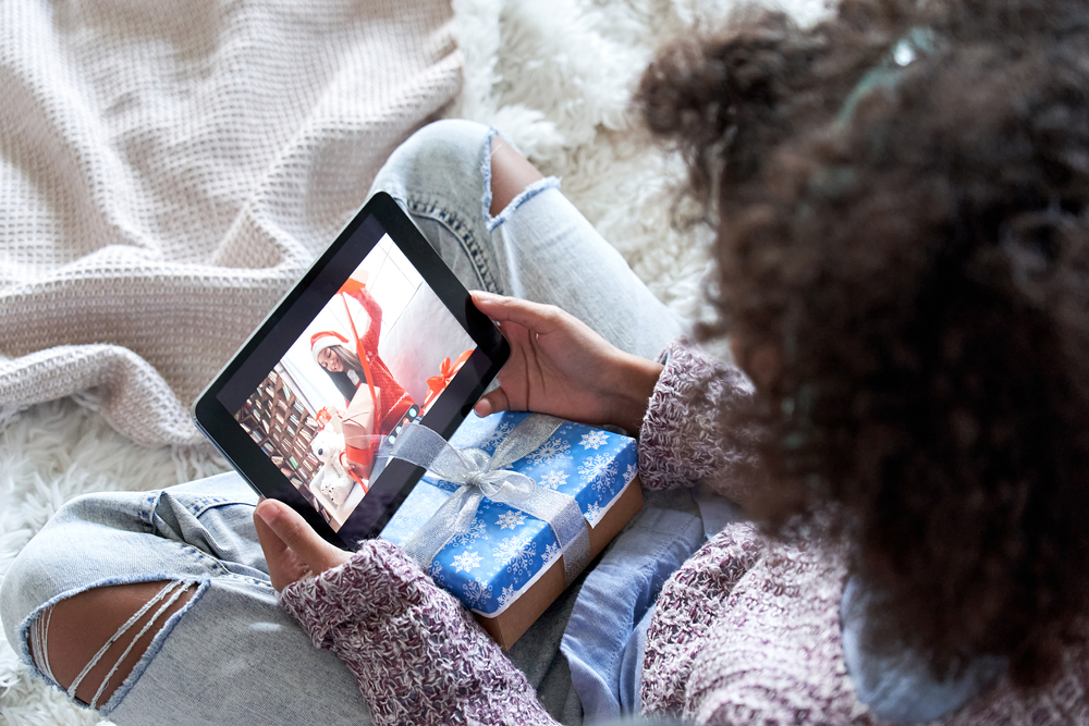 A black girl wearing a knit sweater and blue jeans with a giftwrapped package in her lap holds a tablet, meeting virtually with a friend. 