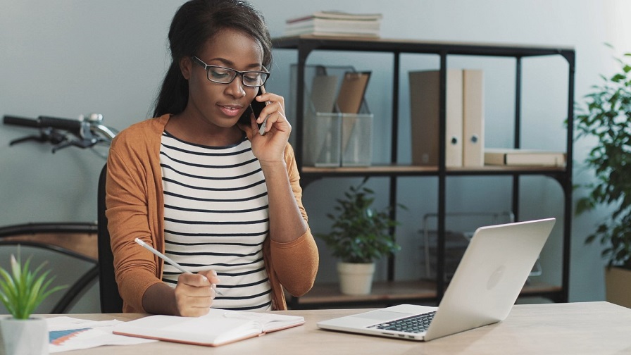 Woman talking into phone, taking notes. A computer is open on a desk.