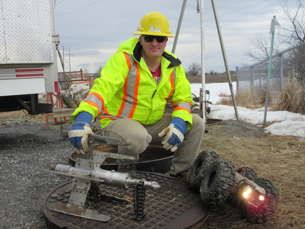 A man in a hard hat, a reflective jacket and work gloves crouches near the access point to a sewer. 