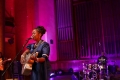 A woman in a blue dress singing at a microphone while playing guitar.