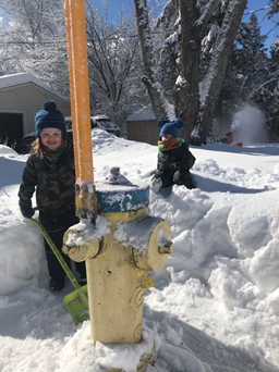 two children dressed in snow gear shoveling snow from around a yellow and blue fire hydrant