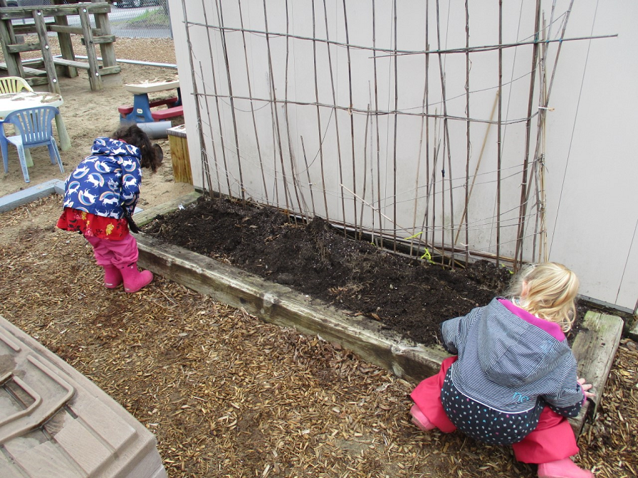 Two kids in spring jackets and rubber boots search for worms in a daycare playground. 