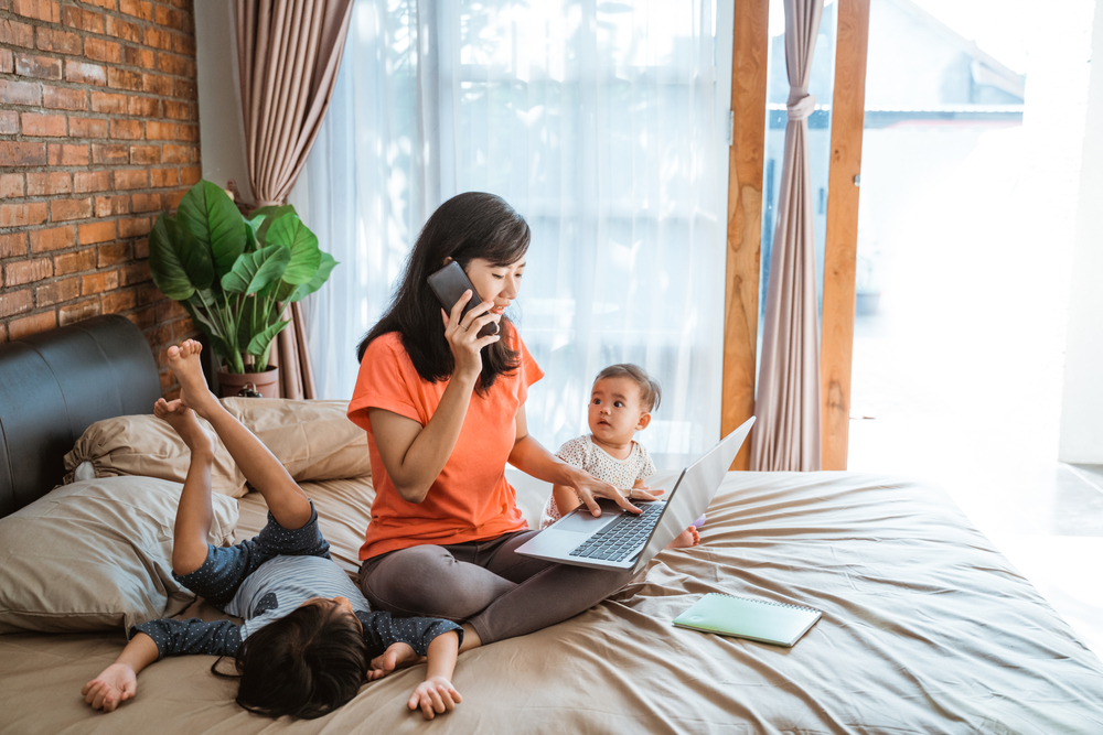 A young Asian mother sitting on a large bed with two young kids, while talking on the phone and looking at a laptop in her lap. 