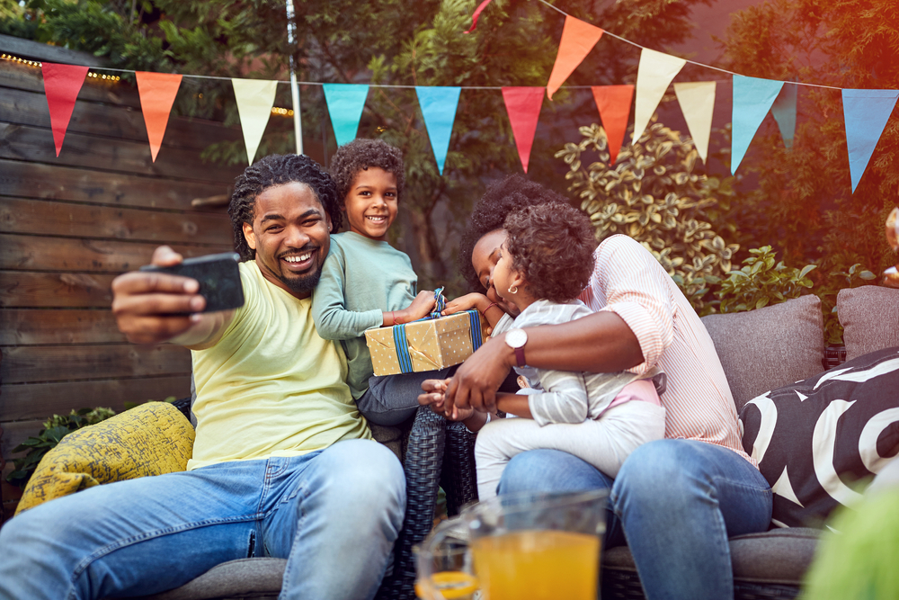 A black man takes a selfie as he celebrates a special occasion outdoors with his wife and two young kids. 