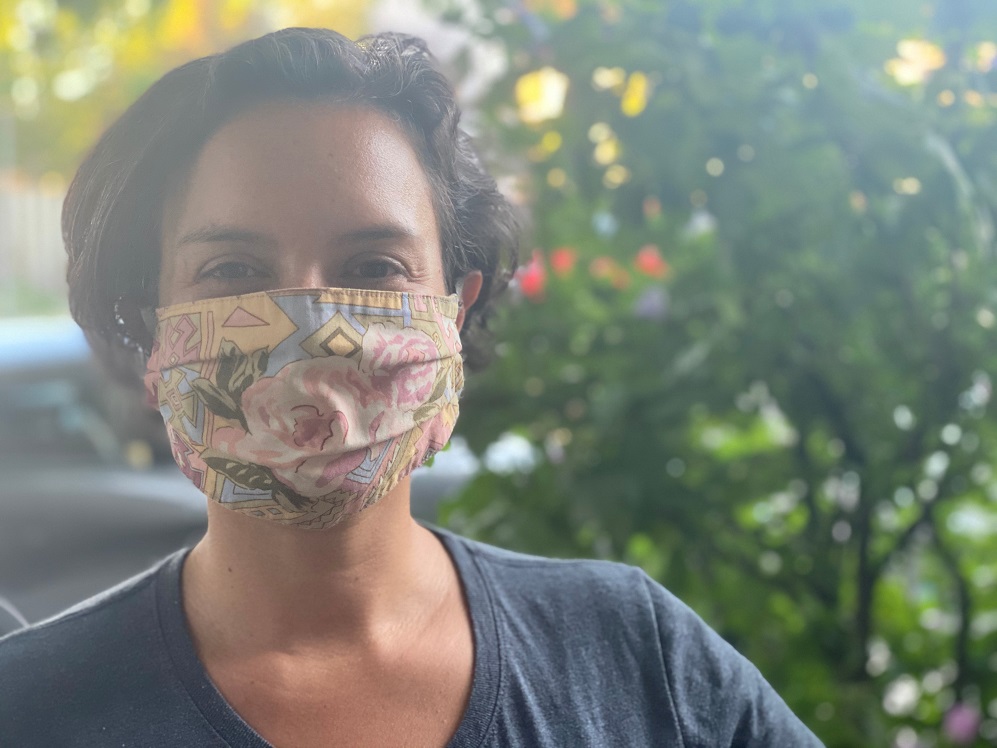 A Hispanic woman wearing a blue shirt and flower-patterned mask stands in front of a tree. 