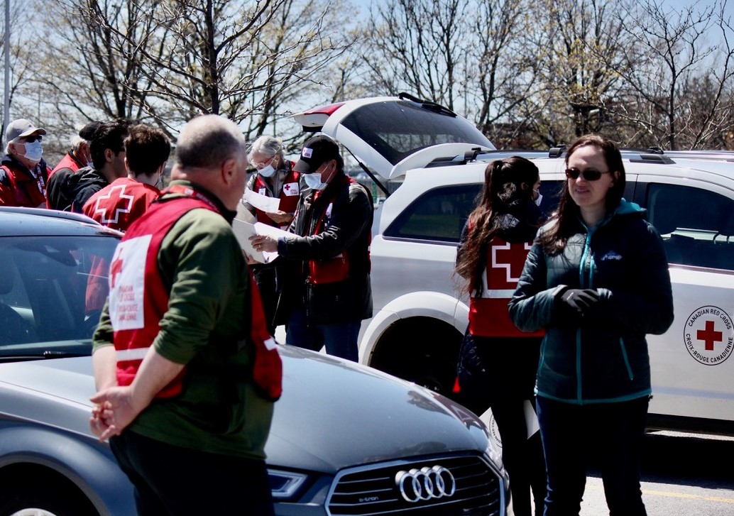 In the background: People gather in a parking lot, wearing masks and red vests. In the foreground, a woman in a dark jacket talks to a man in a green shirt and a red vest. 