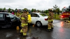 Five firefighters, two damaged cars from a collision, onlookers, and fire truck nearby.